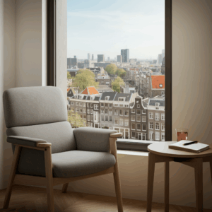 Een moderne grijze fauteuil en bijzettafel bij een groot raam met uitzicht op een levendige Europese stad, passend bij het thema Innerlijke Rust in Amsterdam.