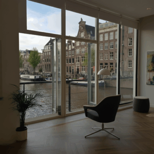 Een stijlvolle therapieruimte in Amsterdam met een moderne fauteuil en een plant, uitkijkend over een historische gracht, passend bij het thema Mentale Ruimte in Amsterdam.